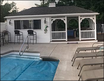 12 x 24 Vinyl Pool House shown with White Siding and Trim, Brown Composite Decking, Driftwood Asphalt Shingles, Cupola, and Concession Window with Bar. Lighting supplied by customer.